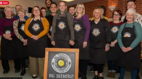 Penerima Big Breakfast Plus berkumpul bersama memakai apron hitam dengan logo Big Breakfast Plus di hadapan, semuanya tersenyum ke kamera