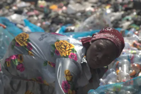 Abiba Alhassan, seorang wanita yang memakai gaun bunga dan bertudung, menyusun botol plastik terpakai di sebelah tapak pelupusan Agbogbloshie di Accra