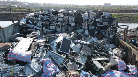 Satu longgokan besar e-sisa, termasuk skrin besar, di tempat pembuangan sampah Agbobloshie. Kelihatan badan air di latar belakang