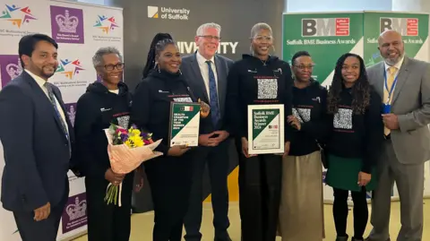 John Wright/BBC Lima wanita dari Sickle Cell Suffolk berdiri dengan sijil anugerah mereka bersama ahli Perkhidmatan Sokongan Pelbagai Budaya BSC dan penaja anugerah Brian Tobin dari Iceni Ipswich