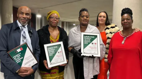 John Wright/BBC Clem Turner, Veronica Akrofi, Beverley Uther, Lydia Forrester dan Angelina Kwamina MBE dari Caribbean dan Forum Sokongan Kesihatan Komuniti Afrika berdiri di dalam Universiti Suffolk yang memegang sijil anugerah.