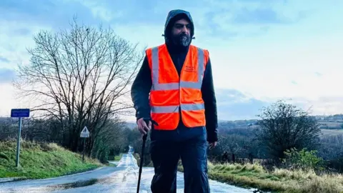 Bhupinder Sandhu Bhupinder Sandhu memakai pakaian kalis air gelap dengan tudung dan jaket pemantul oren. Dia memegang tiang berjalan sambil berjalan di sepanjang jalan desa yang tenang dibarisi pokok dan rumput di laluan antara England dan Scotland. Di latar belakang padang.